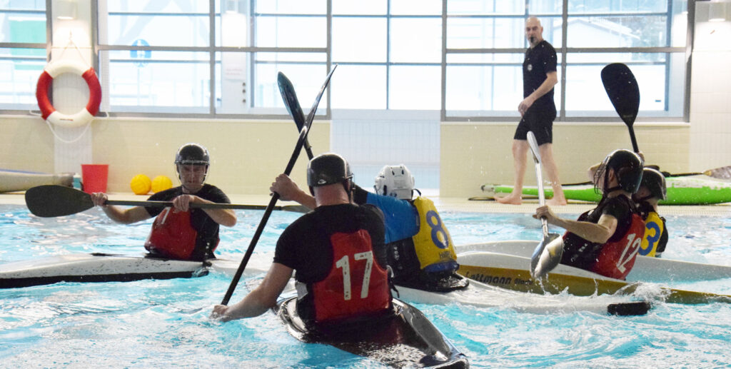 Kanoottipoolokisat. Viisi pelaajaa tavoittelee poolopalloa. Taustalla tuomari seuraa tapahtumaa.